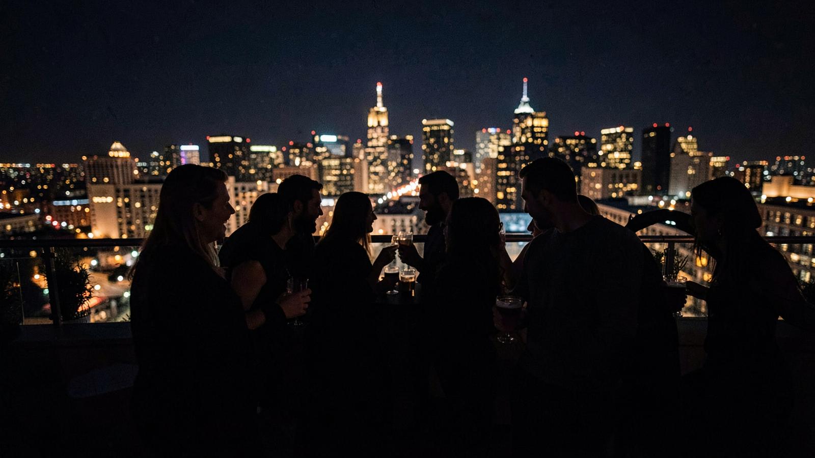 Friends with drinks on a rooftop overlooking a lit city skyline at night